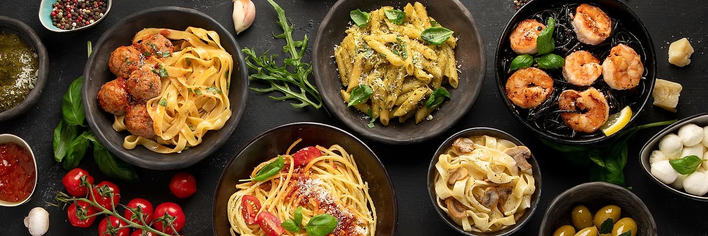 Six bowls of pasta and their garnishes set out across a dark table, shot from above: tagliatelle, penne with pesto, spaghetti, shrimp, olives and mozzarella